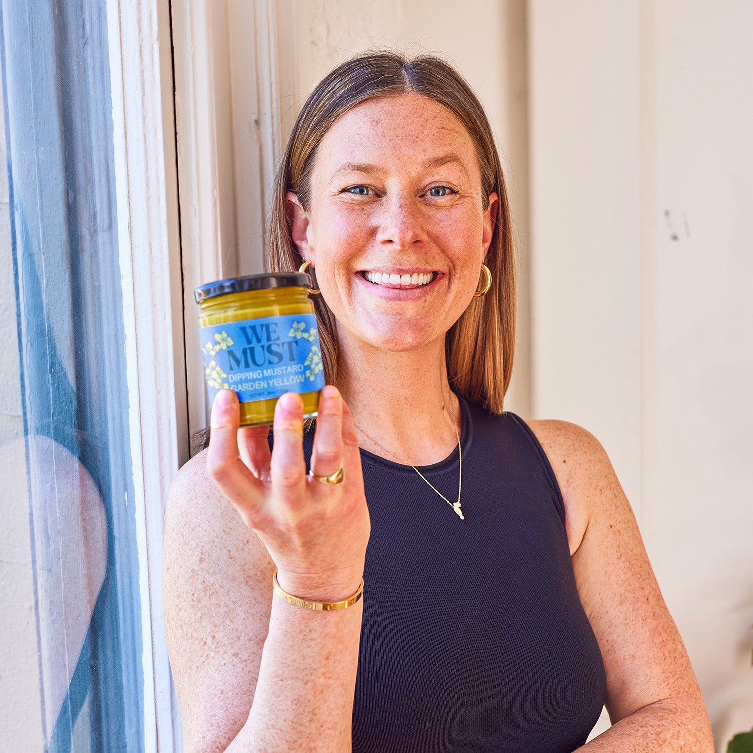 Founder Courtney Yones holding a jar of 'We Must' Garden Yellow Dipping Mustard leaning against a window.