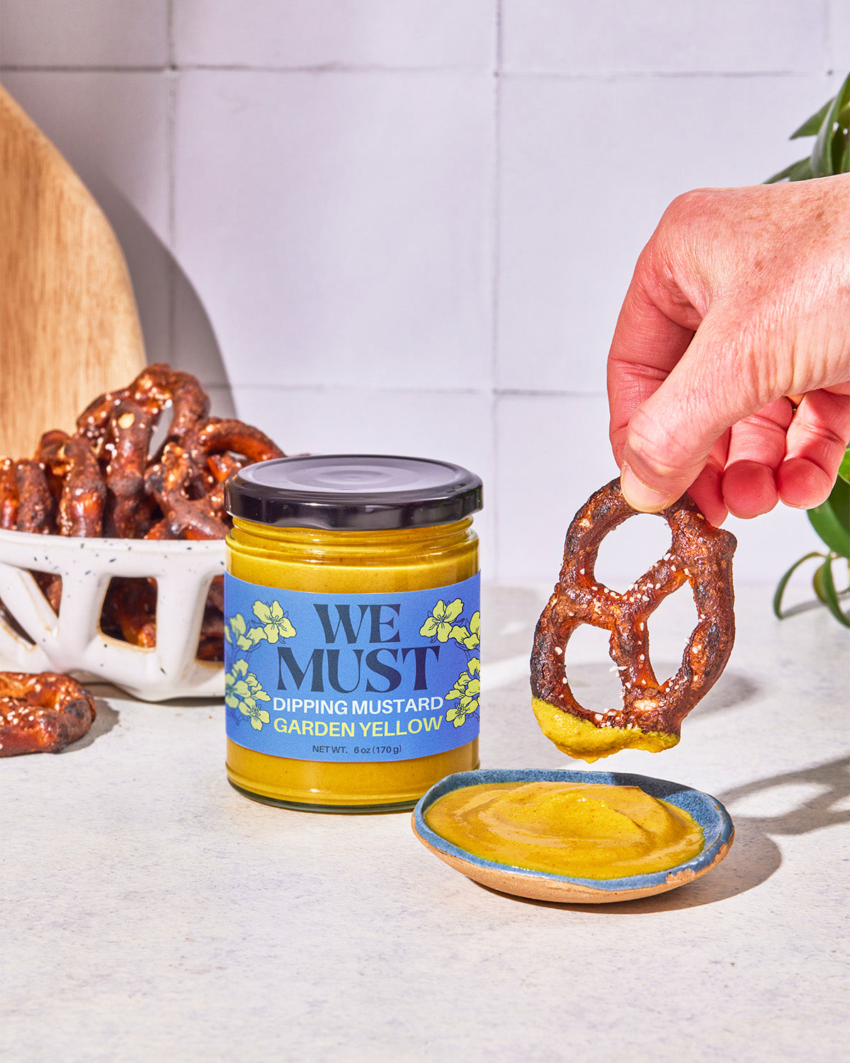 Person dipping a pretzel into a jar of 'We Must" Garden Yellow Dipping Mustard with a visible brand label.
