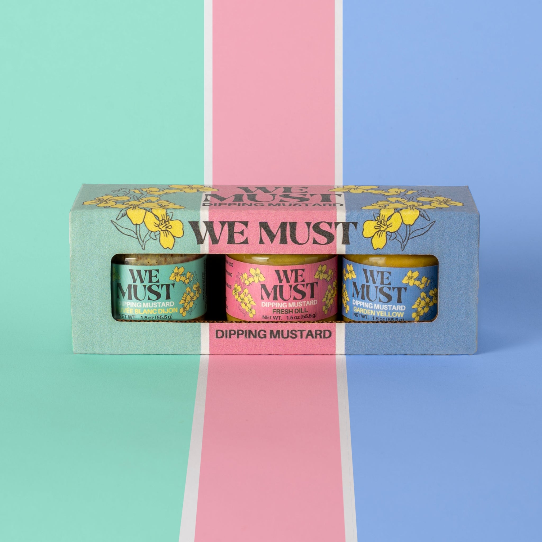 A brightly colored "We Must" Dipping Mustard box containing three 1.5 oz jars of Dipping Mustard. Cuvee Blanc Dipping Mustard, Fresh Dill Dipping Mustard, and Garden Yellow Dipping Mustard. 