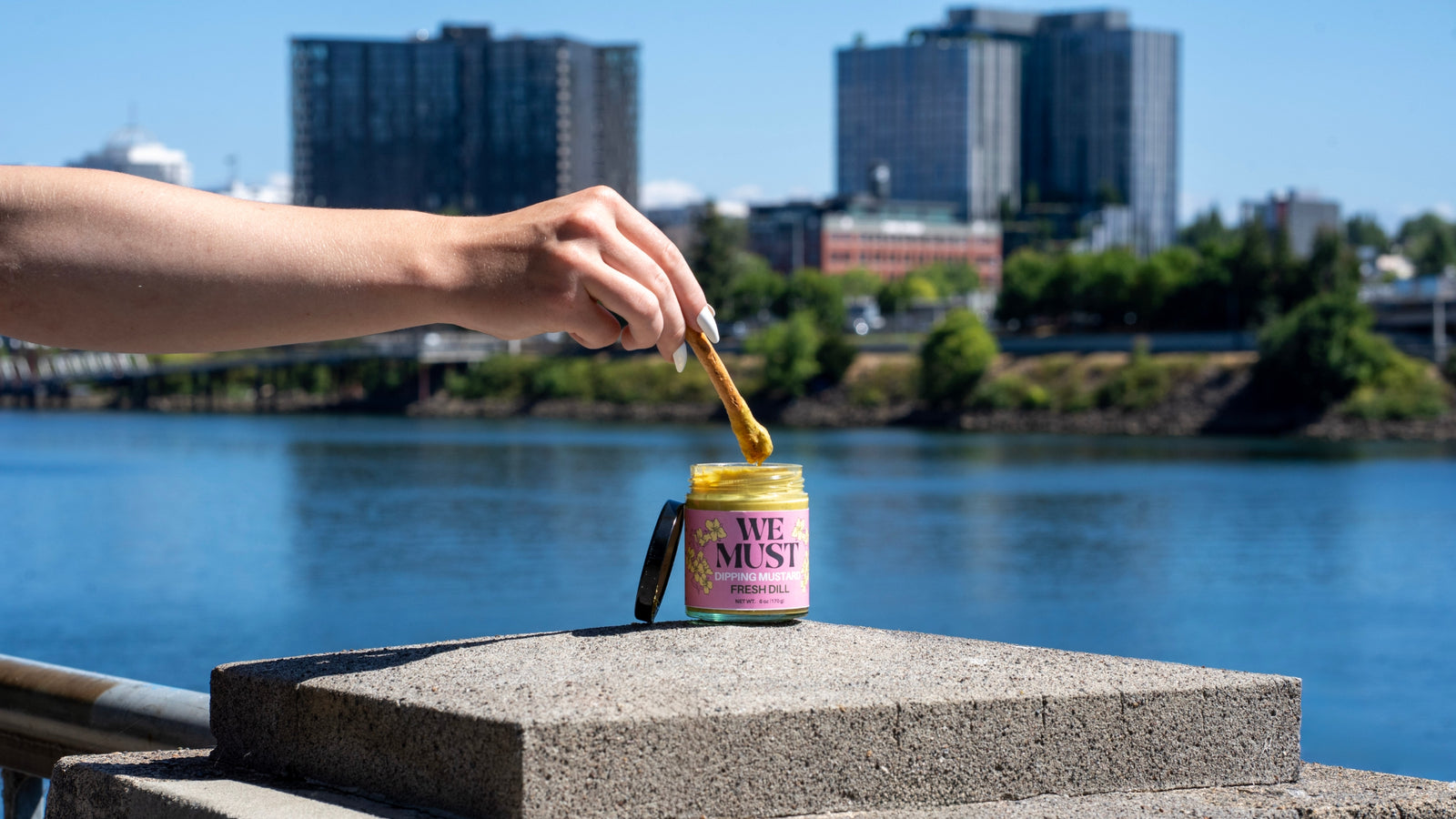 A hand with a white manicure dipping a pretzel stick into a jar of 'We Must' Fresh Dill Dipping Mustard on the Portland Waterfront. 